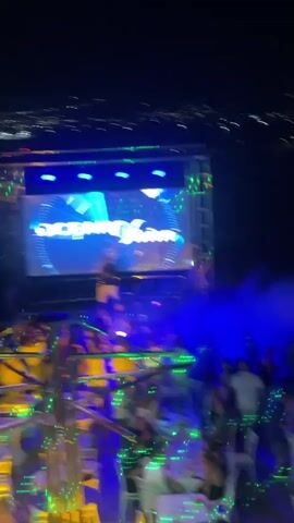 The video shows a nighttime outdoor party with a large LED screen displaying various logos and graphics. The screen displays "SOCBX", "AN6Vision", "OCEANO", and "OCEAN VISION". People are seated at tables and standing around, with some interacting with each other. The lighting is dynamic, with blue and green lights flashing, and smoke effects are visible. The background shows a city skyline at night.