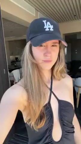The video shows a young woman wearing a black swimsuit with a halter top design and a black LA baseball cap, standing outside on a sunny day. She is looking directly at the camera with a neutral expression. Behind her, there's a glimpse of an outdoor area with furniture and a striped towel. The camera appears to be held by the woman herself, giving a first-person perspective.
