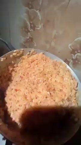 A pot of steaming orange-colored rice is shown up close. The pot looks well-worn. The camera briefly pulls up to show a countertop with outlets and a flower-patterned backsplash. Then, the camera moves to the side, partially blocking the view of the rice.