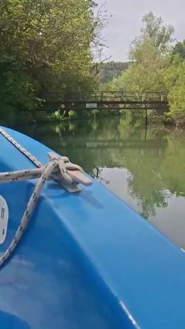 The video captures a first-person view from the bow of a blue boat as it glides along a tranquil, green-hued river. A white rope is coiled and secured at the front of the boat. Visible on the side of the boat are the "facebook" logo and text in Cyrillic, which translates to "eco-trail 'Iskar-Panega'".

The boat approaches a wooden bridge spanning the river. The bridge, with its wooden railings and supports, is surrounded by dense, green foliage and trees on both banks, reflecting clearly in the calm water. A sign attached to the bridge reads "МОСТ НА УСМИВКАТА," translating to "Bridge of Smiles," and several yellow smiley face stickers are visible on its structure. The sky above the trees appears overcast or partly cloudy. The scene depicts a serene boat journey through a natural landscape, characteristic of an eco-trail, likely in the Petrevene region of Bulgaria.
