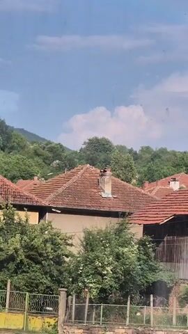 The video captures a view of several tiled-roof houses in a rural setting with a bright sky and fluffy clouds. Lush green trees and vegetation surround the buildings. In the foreground, a fence runs along the bottom of the frame, with a stone wall and some wire fencing visible. The houses appear to be older, with one building prominently featuring exposed brickwork and wooden beams, and another with a prominent chimney. Some parts of the buildings show signs of age or disrepair. The overall impression is of a tranquil, somewhat rustic village.