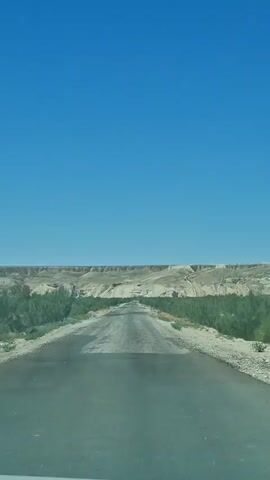 A vertical shot of a paved road stretching into a desert landscape under a clear blue sky. The road appears somewhat weathered and uneven. To the sides of the road are sparse green shrubs and dry, sandy ground. In the distance, rolling hills and rock formations with distinctive erosion patterns are visible. The sun appears to be shining brightly, casting minimal shadows. No people or vehicles are visible.