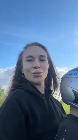 A woman with long brown hair, wearing a black hooded sweatshirt, is recording a selfie video. She is smiling and looking directly at the camera. To her right, partially visible, is a person, likely a child, wearing a silver helmet with pink goggles. The background shows green grass, trees, and a blue sky with some white clouds, indicating an outdoor setting. The scene appears to be in motion, suggesting they are on a moving vehicle, with the woman's hair blowing slightly in the wind.
