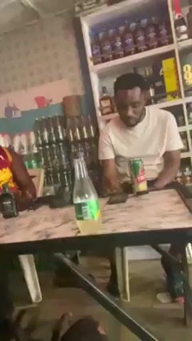 The scene takes place indoors, seemingly inside a bar or a similar establishment. Two men are seated at a table with a marble-like pattern. The man on the left is wearing a Cleveland Cavaliers jersey, and the man on the right is wearing a light-colored t-shirt.

On the table, there is a bottle of Sprite, a can of Club Shandy, and what appears to be a small bottle of liquor. The man on the right is looking at his phone.

Behind the men, there is a shelf stocked with bottles, mostly alcohol, along with some other items like cleaning supplies. The background is a tiled wall. The lighting appears to be somewhat dim and possibly artificially lit. Music is playing in the background.