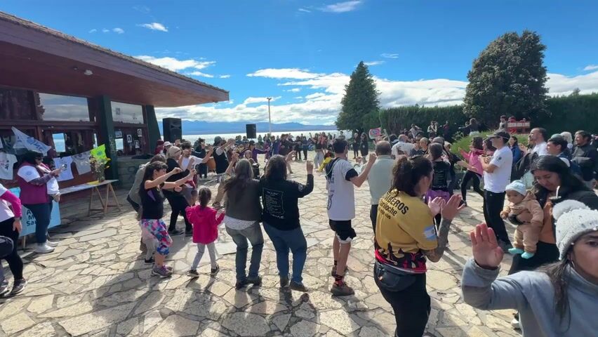 A wide-angle shot captures a large group of people dancing in a public area on a sunny day with a bright blue sky dotted with fluffy white clouds. The dance is taking place on a stone-tiled plaza adjacent to a building with a wooden roof and glass windows, set against a backdrop of a wide lake or sea and distant mountains. Tall green trees stand on the right side of the frame.

The crowd is diverse, with people of all ages, including children, and various ethnicities participating in the dance. They are dressed in casual clothing, including t-shirts, jeans, and jackets, with a few wearing hats or beanies. Some individuals are holding small children or babies. A few people stand on the edge of the crowd, watching the dancers.

On the left side of the frame, a small table is set up with flyers or signs, indicating a possible event or gathering. The scene suggests a community event or celebration.