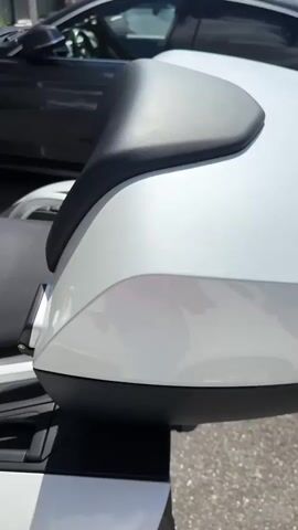 The video begins with a close-up of a white motorcycle seat. The camera then pans to reveal the front of the motorcycle, including the handlebars, windshield, and a mounted screen. A dark-colored car is visible in the background. The motorcycle appears to be parked outdoors, possibly near a building with windows and signage. The video continues with a closer view of the motorcycle's handlebars and fuel tank, showing the BMW logo. The scene then shifts to a wider shot, showing the motorcycle and the building behind it. A person is partially visible entering the frame on the right side of the video. The environment appears to be a sunny day.