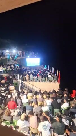 A large crowd of people are gathered outdoors at night, seated on rows of chairs facing a large outdoor screen. The screen appears to be displaying a sports game, possibly soccer, with graphics visible. Many of the attendees are looking at the screen. Flags are displayed on poles behind the screen and to the right. The setting appears to be a tiered outdoor venue, possibly a cafe or stadium, with a railing in the foreground. The lighting is dim, with the screen being the primary light source.