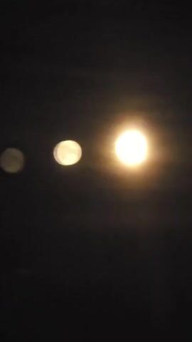 The video shows a dark sky with three blurry circular objects that appear to be lights. The largest and brightest light is on the right, with a smaller, fainter light to its left, and an even fainter, blurrier light to the far left. The lights are arranged horizontally and are slightly out of focus. The background is dark and appears to be night. There are no people or discernible objects visible other than the lights.