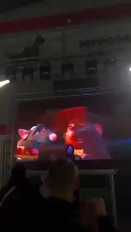 The video shows a large screen in a dark indoor space, initially lit with blue stage lights. The screen displays an animated scene of two cartoon rats interacting. As the camera pans up, more stage lights are visible above the screen, along with a sign reading "HIPPODROM." The camera focuses on the screen, then moves to show the back of a person's head standing in front of the screen. White stage lights shine directly onto the screen and person.