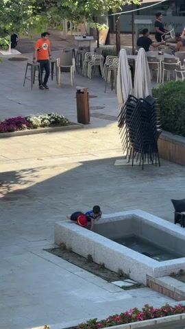 A young child wearing a blue shirt, red shorts, and a red hat is lying on their stomach on the edge of a rectangular concrete fountain. The child is looking down into the fountain. Another child, wearing a red shirt and blue shorts, is lying next to the first child and also looking into the fountain. The fountain appears to contain water. In the background, there are stacks of chairs, tables, umbrellas, and people sitting at tables outside a building, suggesting an outdoor cafe or restaurant setting. A person in an orange t-shirt is walking in the middle ground. The ground is paved. Trees are visible in the upper left of the frame.
