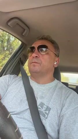 A middle-aged man with short, grey hair and sunglasses is driving a car. He is wearing a grey t-shirt with a skull and crossbones logo on it. He has a seatbelt on. He is wearing wireless earbuds in his right ear. The interior of the car is visible, including the steering wheel, dashboard, and the passenger side door frame. Outside the car, trees and foliage can be seen. The video is shot from a first-person perspective, looking forward from the driver's seat.