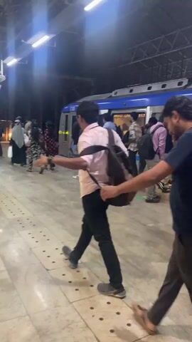 At a subway station, several people are disembarking from a blue and silver train, visible behind them. Passengers with backpacks walk away from the train, while others exit through the open doors. The station platform is illuminated by bright overhead lights, and the ceiling is high and angled. The floor is tiled with a pattern of off-white dots.