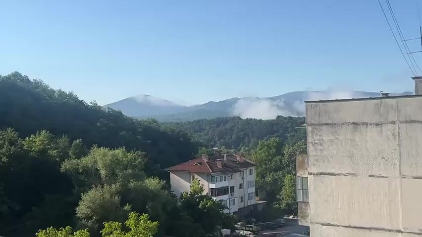 The video pans from right to left showing a hazy mountain range in the distance, covered in low-lying clouds. In the foreground are several apartment buildings, some appearing older and weathered, others more modern. Lush green trees surround the buildings, and the sky is a clear blue. The scene appears to be a residential area nestled against a natural landscape.