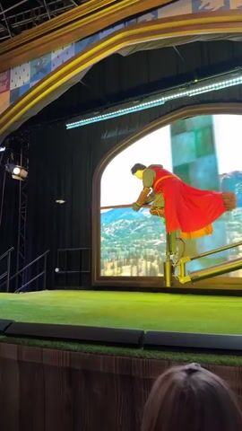 This video shows a performer dressed as a Quidditch player on a broomstick, suspended in mid-air on a mechanical rig. The player is wearing a red robe over a green and brown uniform, with gloves and goggles. They are positioned in front of a large screen displaying a scenic mountainous landscape with a blurred effect, suggesting rapid movement. The stage has a green grassy floor and a wooden platform in the foreground. The background features a curved archway with decorative panels and a black curtain. Lights are visible above the screen and on a stand to the left. The scene appears to be part of a live performance or attraction.