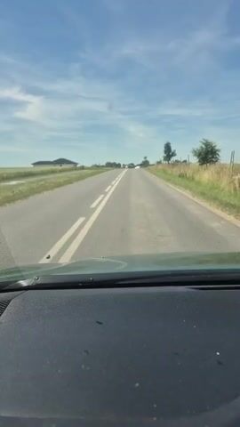 The video shows a first-person perspective of a car driving down a paved road in a rural setting. The road is in good condition with clear white lane markings. The surrounding landscape consists of grassy fields and scattered trees, with a few houses and buildings visible in the distance. The sky is blue with white clouds, suggesting a clear and sunny day. The time of day appears to be daytime, likely late morning or afternoon, given the brightness and the shadows.

There are no people visible in the video. The primary objects are the car's dashboard in the foreground and the road and landscape outside the car. The scene depicts a calm and uneventful drive through the countryside. There are no overt emotional cues, but the steady forward motion and open road can evoke a sense of freedom or tranquility.

There is a traffic sign visible on the right side of the road around the 00:02 mark. It is a circular sign with a red border and a black symbol inside, indicating a restriction. Based on its appearance, it likely signifies the end of a speed limit or another traffic regulation. There is no other discernible text in the video.

The location context is provided as Mnichowice, Poland, which aligns with the typical rural landscape of Eastern Europe. The overall impression is that of a peaceful journey through a Polish countryside.