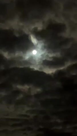 The video shows a full moon peeking through dark, turbulent clouds at night. The moon is partially obscured, casting a faint glow through the atmospheric conditions. The clouds are dense and appear to be moving, creating a sense of motion and drama in the sky. The overall impression is one of a mysterious and somewhat ominous night sky.