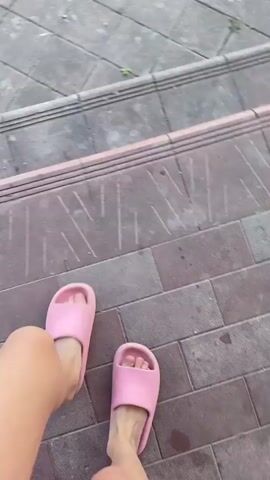 This video captures a person's feet, clad in pink slides, as they walk across a tiled path and then onto a grassy lawn. The camera perspective is from above, looking down at the individual's legs and feet.

The scene is set outdoors, likely in a residential area, given the presence of a brick wall, an ornate metal gate, and houses in the background. The time of day appears to be daytime, with bright sunlight casting shadows. The weather seems to be pleasant, with a clear blue sky and some clouds visible.

The activity is simply walking. The environment suggests a casual setting, possibly a backyard or a pathway leading to a property entrance. The overall mood is relaxed and ordinary, depicting a simple moment of movement. There is no visible text in the video. The location is specified as Murovane, Ukraine.