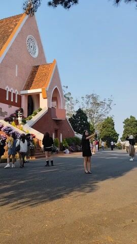 The video shows a woman taking a selfie in front of a pink church. She is wearing a black top, a black skirt, sunglasses, and sandals. The church has a red-tiled roof and is surrounded by trees and flowers. There are other people walking around in the background. The sky is blue and the sun is shining. The ground appears to be paved.