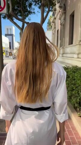 A woman with long blonde hair walks down a sidewalk. She is wearing a white oversized button-down shirt, black lace shorts, and black heeled sandals. A black belt cinches her waist. She walks past a tree and a building with ornate architectural details. A street with cars and a bus is visible on the left side of the frame. The sun casts shadows on the brick sidewalk.