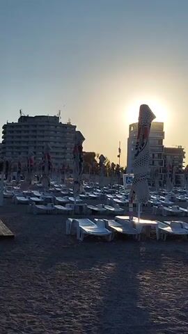 The video shows an empty beach with rows of white lounge chairs and closed umbrellas. The sun is setting or rising, casting a warm glow and long shadows across the sand. In the background, there are several buildings, possibly hotels or resorts. The sky is a clear blue with a hint of orange near the horizon. There are no people visible in the video.