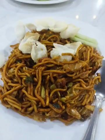 Two plates of food are visible on a white marble table. The plates contain dark brown noodles with bits of vegetables, topped with white chips and slices of cucumber. Each plate has a spoon and fork set next to it. The background is slightly blurred, showing a red chair and possibly other people. The lighting is bright and even, highlighting the texture of the food and the table surface.