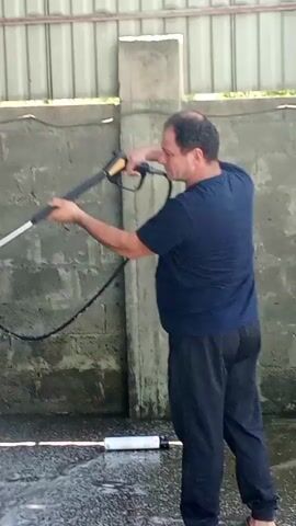 A man in a dark blue t-shirt and black pants is pressure washing a white SUV in a covered area with a concrete wall and a corrugated metal roof. The SUV is covered in white foam and has a spare tire mounted on the back. The man is holding the pressure washer wand with both hands and directing the spray at the car. Water and foam are spraying from the wand and running down the side of the car and onto the ground. There is a black hose connected to the pressure washer. The ground is wet and appears to be concrete.