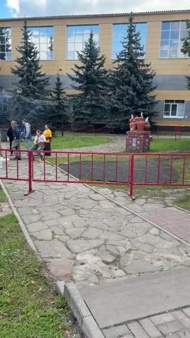 A group of people are gathered outdoors near a building with tall evergreen trees. In the foreground, a red metal barrier divides the scene. A paved walkway runs behind the barrier, and a grassy area is visible to the left. On the right side of the walkway, a decorative sculpture is present. A few individuals are standing and talking, while one person is seated on a chair. The overall atmosphere appears casual and relaxed, possibly during an outdoor event or gathering. The lighting suggests it is daytime, with some clouds visible in the sky.