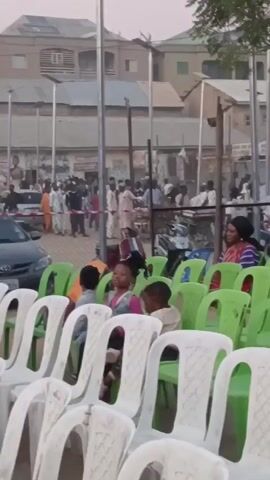 The video shows rows of empty white and green plastic chairs in front of a gathering of people. There are some children sitting in the chairs. Behind the rows of chairs, people are standing and milling around. There are some cars parked behind the chairs. A woman is holding a baby and walking towards the left side of the frame. There are buildings in the background. The sky is overcast. There is music playing in the background.
