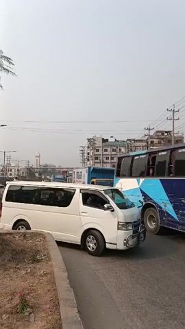 The video shows a busy street in what appears to be a South Asian city. A white van is stopped near the camera, with a blue bus behind it. A yellow truck with a large diaper advertisement passes by. Several cycle rickshaws are present, carrying passengers, and people are walking along the side of the road and crossing the street.  In the background, there is a pedestrian bridge, buildings, and a mosque minaret visible. The sky is overcast and hazy.
