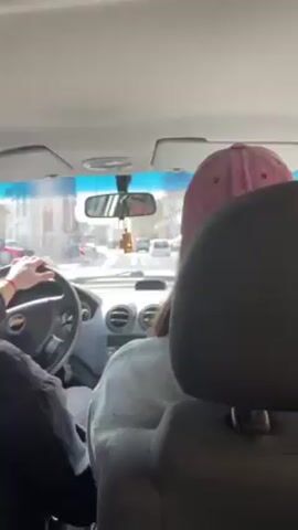 The video is shot from the back seat of a car. A person wearing a black jacket drives, with a hand visible on the steering wheel. Another person in the passenger seat wears a light gray top and a pink baseball cap with lettering on it. The car is moving along a street with buildings visible on the side. There are other cars ahead. The interior of the car is visible, including the dashboard and rearview mirror.