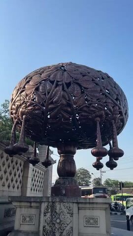 A copper-colored, ornate, dome-shaped sculpture with a leaf motif rests atop a pedestal. The sculpture has decorative tassels hanging from its underside. The pedestal is made of stone and features carved embellishments. To the left of the sculpture is a light-colored stone wall with a repeating diamond pattern. In the background, there is a clear blue sky, green trees, and a street with cars and trucks. The scene appears to be outdoors during daylight.