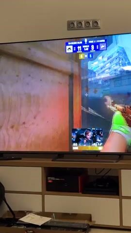 A television screen displays a first-person shooter video game. The game features a colorful, urban environment with buildings and vehicles. In the foreground, a player's virtual hands are holding a gun with a green and red patterned skin. The player is aiming at a yellow truck that is engulfed in flames and smoke, with purple and pink explosions emanating from it. A smaller window in the bottom right corner of the screen shows a live feed of a man, identified as "MAX," playing the game, with his face illuminated by the screen's light. The game's interface at the top of the screen shows team scores, round information, and timers. The television is placed on a white media console with black electronic devices underneath. The room appears to be dimly lit.