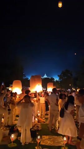 At night, a large group of people dressed in white are gathered in a grassy area to release sky lanterns. The sky is dark blue, and numerous lanterns are floating upward, glowing warmly against the night. In the distance, a bright, illuminated structure can be seen through the trees. Closer to the foreground, people are clustered together, some holding up lanterns that are lit and ready to be released.  Small candles on stands are placed throughout the area. The atmosphere is festive and peaceful.