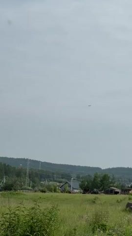An airplane flies high in the sky over a rural landscape. Below the airplane, power lines stretch across the frame, supported by tall metal towers. In the foreground, a grassy field is visible, with trees and houses scattered throughout the middle ground. The sky is overcast, with the sun not clearly visible.
