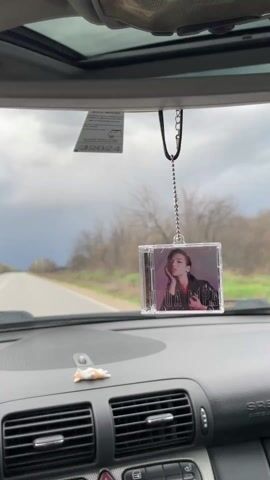The video shows a driver's perspective from inside a car, focused on a road ahead. A clear plastic CD case with a Dua Lipa album cover hangs from the rearview mirror. The dashboard of the car is visible, featuring air vents, hazard lights, and other controls. A small white object sits on the dashboard. Outside, the road stretches into the distance under a dramatically cloudy sky, with trees and sparse vegetation lining the sides. The car is moving.