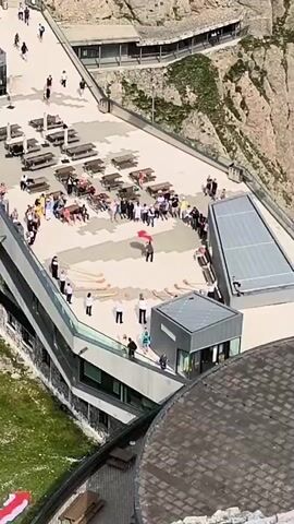 The video shows a bird's-eye view of a rooftop with many people gathered. The rooftop appears to be part of a modern building with large windows and a balcony area. There are several picnic tables with umbrellas spread across the rooftop, some occupied by groups of people. A large red flag with a white star is being unfurled by several individuals in the center of the rooftop. The surrounding environment is mountainous with steep, rocky cliffs and some vegetation. Another building with a dark, textured roof is visible in the foreground. The sky is bright, suggesting it is daytime.