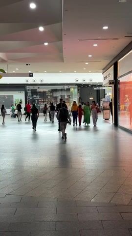 The video shows an indoor scene within a modern shopping mall located in Ciudad Guayana, Venezuela. Numerous people, including adults and children, are seen walking along a wide, tiled concourse. The mall features high ceilings with recessed lighting and several storefronts with large glass windows displaying merchandise. On the right side, a storefront displays a "20%" sale sign, and a partially visible sign suggests a brand like "Michael Kors" or similar. Further down the corridor, another sign reads "FERIA OK." In the background, an arrow sign points upwards, likely indicating another level or specific area within the mall. The overall atmosphere is bustling but calm, with individuals and groups moving leisurely through the shopping area. The lighting is bright and artificial, typical of an indoor commercial space.