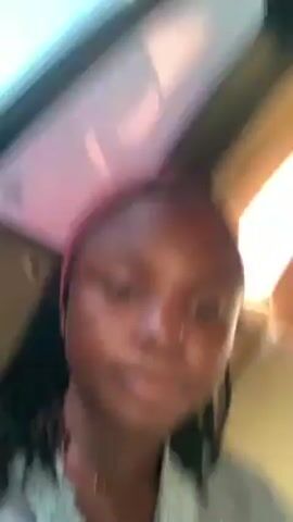 The video shows a close-up of a person with dark skin and braided hair in an indoor setting. The person is wearing a gray garment with white lace trim and a delicate gold necklace. They appear to be looking directly at the camera. The background is blurry, suggesting the person is in motion or the camera is unstable. Light is coming from an overhead source.