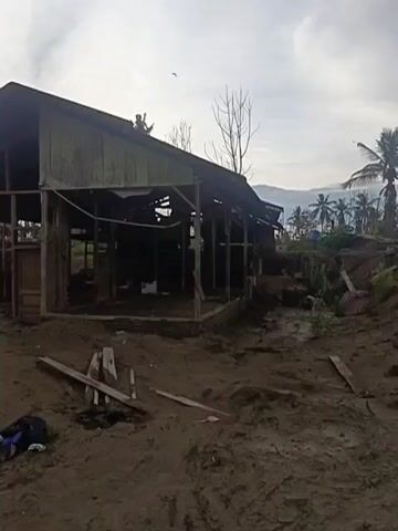 The video shows a close-up of a damaged wooden structure with a corrugated metal roof. The structure appears to be partially dismantled, with exposed wooden beams and no walls. The ground around the structure is covered in dirt and debris, including wooden planks and a blue bag. In the background, more similar structures can be seen, some with metal roofs and others under construction. Palm trees and a cloudy sky are visible in the distance. The overall impression is one of damage and disrepair, possibly due to a natural disaster or ongoing construction.
