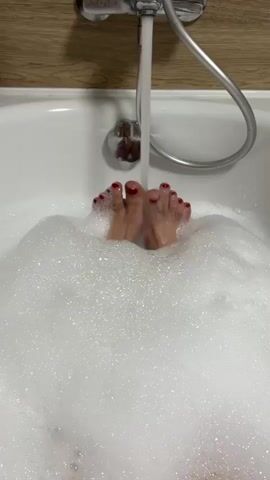 The video shows a close-up view of a person's feet submerged in a bathtub filled with white foam. A stream of water is being poured from a faucet onto the feet. The feet have red nail polish on the toenails. The bathtub appears to be white and is set against a background of a wooden-patterned wall. The water level rises as the foam bubbles around the feet.
