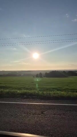 This video shows a view from inside a vehicle moving along a paved road. The sun is high in the sky, casting a bright glare and lens flares. In the distance, there is a vast green field bordered by a treeline. A metal pole with a sign is visible on the left side of the road. Power lines stretch across the sky. The scene suggests a sunny day in a rural or agricultural area.