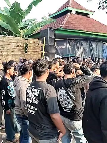 The video shows a large crowd of people gathered in an outdoor space that appears to be muddy. Most of the individuals are wearing dark clothing, some with printed designs on their shirts. A building with a red roof is visible in the background, along with trees and banana plants. Some people are moving or dancing, with their arms raised, while others are standing and watching. A speaker system is set up near the building. The camera pans to show the extent of the crowd and the surrounding environment.  Some people hold what appear to be whips.