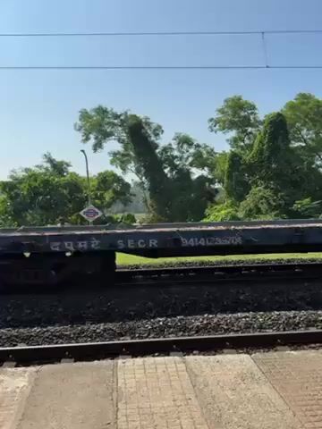 The video shows a flatbed rail car moving on a track. The car is mostly blue, with some rust. The video is taken from a slightly elevated position, possibly a platform. Trees and some buildings are visible in the background under a clear blue sky. The focus is on the rail car as it passes by.