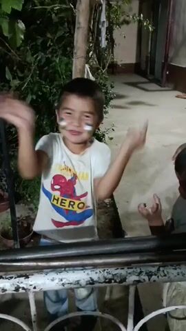 The video shows two young boys playing outdoors in what appears to be a residential area. One boy, wearing a white t-shirt with a Spider-Man "HERO" graphic and plaid shorts, has face paint on his cheeks in green and blue, resembling a flag or design. He is smiling and animated, with his arms outstretched as if dancing or playing a game. The second boy, in a white t-shirt, is partially visible to the right, also engaged in the interaction.

The setting is somewhat rustic, with leafy green foliage visible behind the first boy, suggesting a garden or natural area. A wooden pole and some white cloth are also present. In the background, a paved walkway or patio area is visible, leading towards a building with a doorway. The lighting suggests it is either dusk or nighttime, with artificial light sources illuminating the scene.

The children appear to be having fun and are actively interacting with each other, possibly playing a game of tag or a similar energetic activity. The overall mood is playful and joyful. The visible text on the first boy's shirt reads "HERO" in yellow letters above a red Spider-Man figure. The time stamp on the video indicates it was recorded in 0.00 to 0.05 seconds, suggesting a very short clip. The location context provided is Erkin, Uzbekistan.
