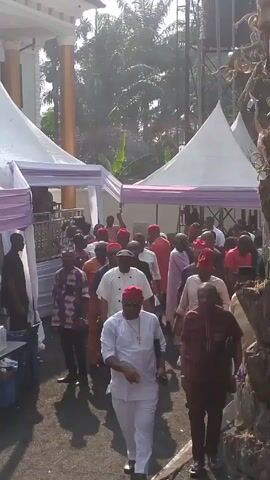 The video shows a large group of men walking outdoors, likely at an event.  Most of the men are wearing red hats.  They are walking between white tents with light purple trim.  Some men are wearing traditional African attire.  Others are in more modern clothing, like button-down shirts or suits.  The background includes a building with tall columns and trees.  The lighting is bright, but there is some glare from the sun.  There is a table to the left with water bottles on it.