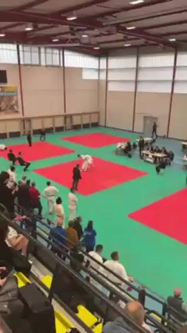 The video shows two people in white judo uniforms competing on a red and green mat. One person throws the other, and they both end up on the mat. A referee in a black suit stands nearby, watching the competition. Several spectators are visible around the mat. There is a table with people sitting at it and a digital scoreboard showing the names "BRUNSTEIN M" and "PARGALAVA N" with scores of 22 and 11, respectively. The weight class is listed as "90 kg," and the timer shows "0:30." The environment appears to be an indoor sports hall.