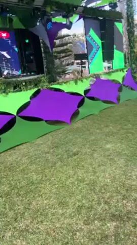 The video shows a DJ performing at an outdoor event. The DJ is positioned on a stage decorated with green foliage and digital screens displaying geometric patterns. The stage is also lit with blue and green spotlights. In front of the stage is a seating area with purple and green diamond-shaped canopies over a grassy lawn. The DJ appears to be operating a mixing console and interacting with the audience. The atmosphere seems festive, suggesting a music festival or outdoor party.