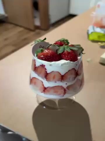 A video shows a dessert consisting of layers of strawberries and white cream in a stemmed glass. The top of the dessert is decorated with three whole strawberries and a small label. The dessert sits on a wooden surface with a refrigerator in the background. A person's hand briefly touches the glass. The person in the video mentions that their landlord gave them the dessert, and that it is a homemade one.