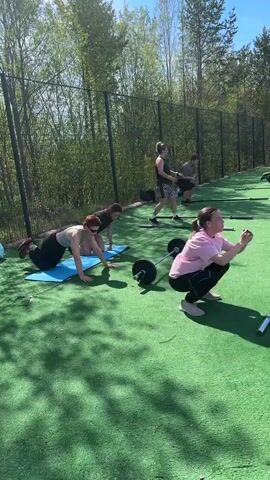 The video shows an outdoor workout scene. Four women and one man are visible on a bright, sunny day. The setting appears to be a training area with artificial green turf, surrounded by a wire fence and trees. In the foreground, a woman in a pink shirt and black pants is doing squats. Next to her, a woman with short red hair is doing push-ups on a blue exercise mat. A barbell lies nearby. In the background, another woman is standing with her hands on her hips, and a man is doing arm exercises. A third woman stands nearby. The scene suggests a group fitness session or a personal training session.