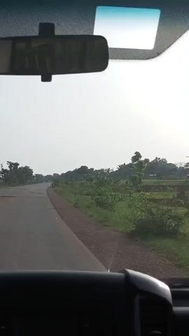 The video shows a car driving on a paved road. The road is bordered by grass and trees. The sky is overcast. The rear view mirror and a blue visor are visible through the car’s front windshield.